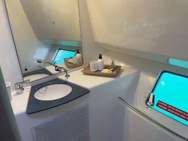 This image shows a compact, modern bathroom with a triangular sink, large mirror, and toiletries on a tray. A window reveals underwater views with turquoise lighting.