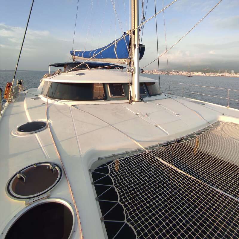 The image depicts a white catamaran sailboat with a blue sail cover, floating on the water, with a city skyline visible in the background.