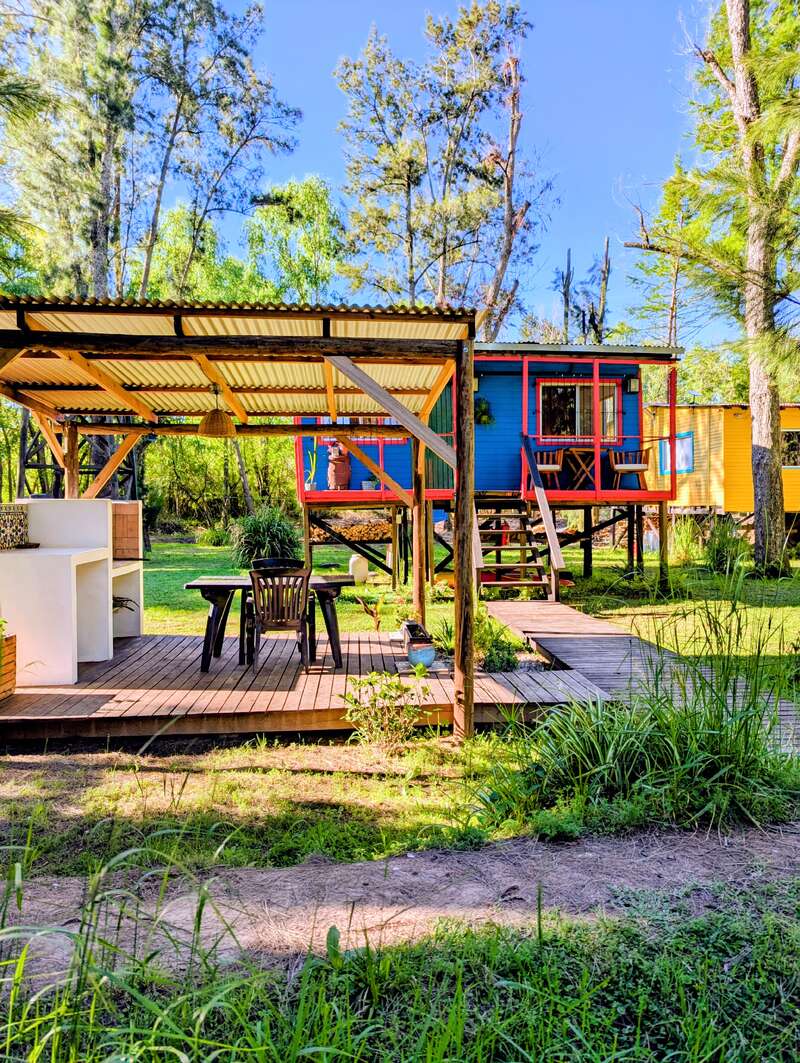 Eine gemütliche Holzterrasse mit Tisch und Stühlen steht neben einem knallblauen Tiny House, umgeben von hohen grünen Bäumen unter einem strahlend blauen Himmel.