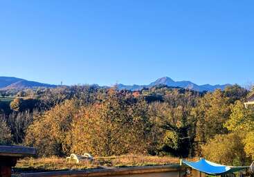 Un pintoresco paisaje otoñal con árboles llenos de color, montañas lejanas bajo un cielo azul y despejado, y algunos muebles de jardín en primer plano. La escena irradia tranquilidad y belleza.