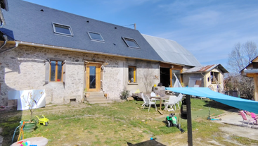 Una casa de piedra con techo de pizarra se alza bajo un cielo azul brillante. En el patio hay muebles de exterior, juguetes infantiles y diversos objetos esparcidos por todas partes.