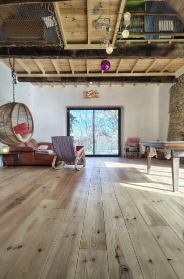 Un salón acogedor y rústico con suelos y techo de madera, una silla colgante de ratán, un sofá, una mecedora, una mesa de billar, un gran ventanal y luz natural. Ambiente acogedor.