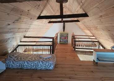 Esta imagen muestra un acogedor loft en un ático con paredes y suelo de madera, un colchón a la izquierda, estanterías al fondo y barandillas de estilo rústico.