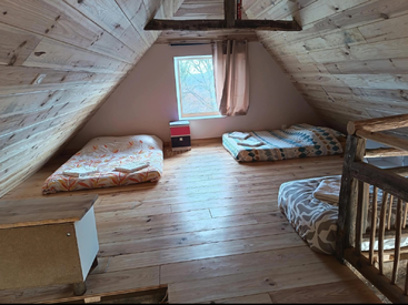 Se trata de una acogedora habitación en el ático con paredes y suelos de madera, tres colchones en el suelo, una ventana con cortinas, una barandilla rústica y una decoración sencilla y minimalista.