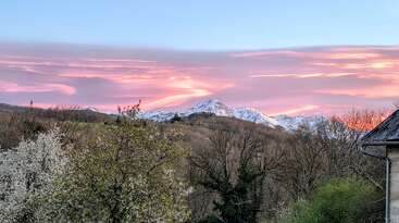 Un paisaje impresionante en el que se ven montañas nevadas bajo un espectacular cielo anaranjado y púrpura al atardecer, enmarcado por árboles en flor y un edificio rústico en primer plano.