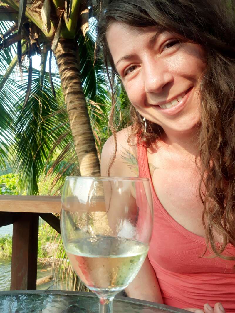 La imagen muestra a una mujer de pelo largo y castaño, con una camiseta rosa de tirantes, sentada a una mesa con un vaso de agua delante, rodeada de palmeras.