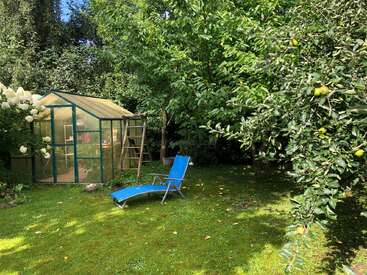 Une scène de jardin paisible avec une serre en verre, une chaise longue bleue, des arbres fruitiers, de l'herbe verte luxuriante, des fleurs épanouies et la lumière douce du soleil filtrant à travers les feuilles.