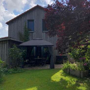 Cette image montre une maison en bois avec un jardin. Il y a une gloriette avec des meubles d'extérieur, une végétation luxuriante, un arbre à feuilles rouges et une jardinière. Atmosphère paisible.