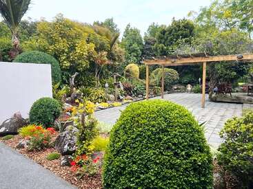 A lush, vibrant garden with trimmed bushes, flowering plants, stone pathways, a wooden pergola, and a seating area. Trees and greenery surround the serene space.