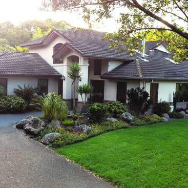 A modern, single-story house with a tiled roof, surrounded by lush landscaping, green lawn, tropical plants, trees, rocks, and a winding driveway, exuding tranquility and elegance.