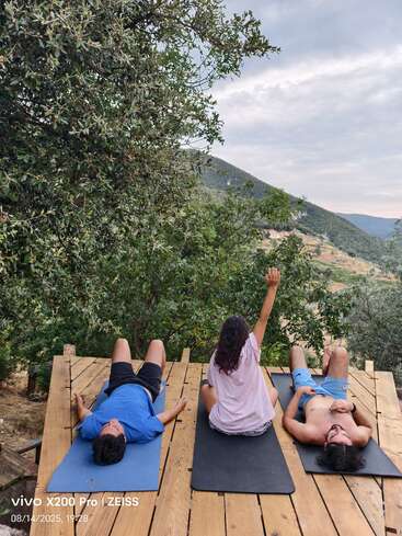 Three people relax on yoga mats atop a wooden deck, surrounded by lush trees and hills. One person sits upright, arm raised toward the cloudy sky.