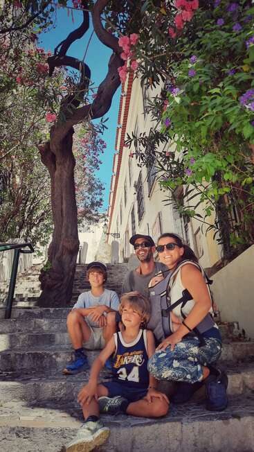 Una familia feliz posa en unos escalones de piedra rodeada de florecientes flores rosas y moradas, bajo un árbol nudoso, disfrutando de un día soleado en un pueblo encantador.