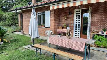 Una acogedora casa de ladrillo con patio cubierto, mantel a cuadros sobre una mesa de madera, bancos, plantas de jardín, sombrilla y un ambiente que invita a reunirse al aire libre.
