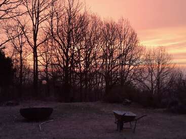 La silueta de unos árboles desnudos se recorta contra el cielo rosáceo anaranjado del atardecer. En primer plano, una carretilla y una hoguera descansan sobre el suelo helado. Un atardecer tranquilo.