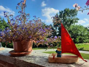 Una maceta con flores moradas descansa sobre una repisa de piedra junto a una figurita de velero con una vela roja, bajo un soleado cielo azul.