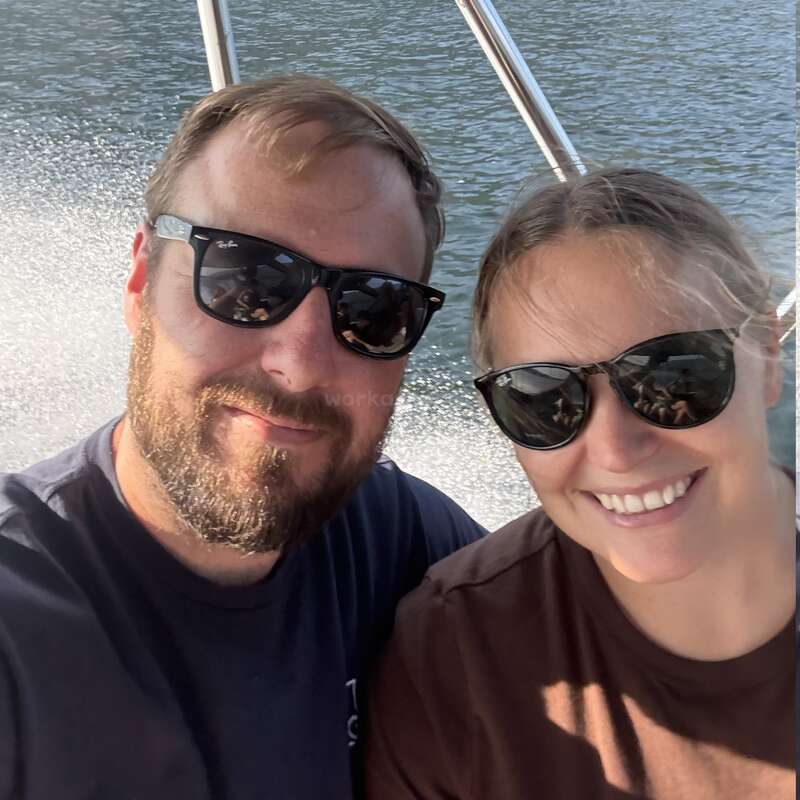 A smiling man and woman wear sunglasses while sitting on a boat. The sun is shining, water sparkles behind them, and they appear to enjoy the ride.