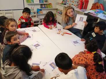 The image shows a woman teaching a group of children at a table, with papers and pictures of people on them, in a classroom setting with bookshelves.