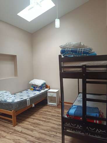 This room features a skylight, wooden flooring, a single bed, bunk bed, white nightstand, and several folded blankets and pillows, creating a basic, cozy space.