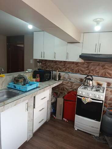 This image shows a small kitchen with white cabinets, a gas stove, blender, kettle, dish rack, microwave, red trash bin, and patterned backsplash, featuring wood flooring.