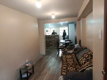 This image shows a cozy living room with patterned sofas, a small table, wooden flooring, dining table, and two people standing in the kitchen area.