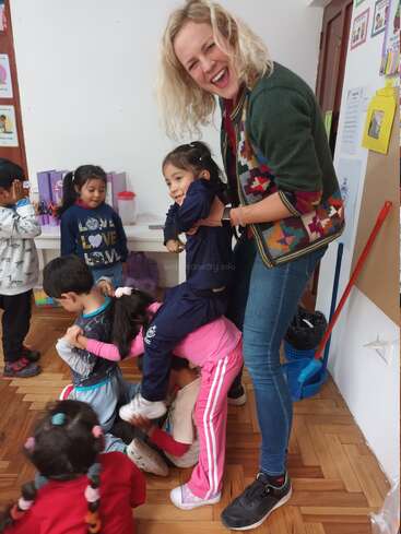 A joyful woman laughs as several young children hug her and play together in a colorful classroom, creating a warm, happy, and playful atmosphere filled with smiles.