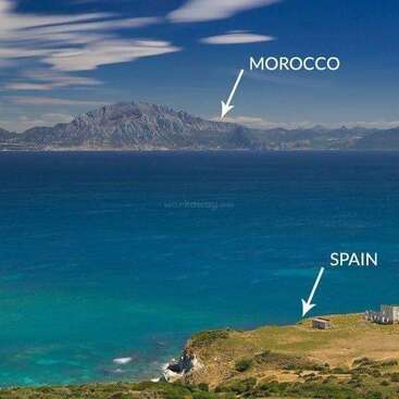 The image depicts a serene coastal scene with a mountainous backdrop, featuring a body of water and a grassy cliff, with the labels \"MOROCCO\" and \"SPAIN\" indicating the location.
