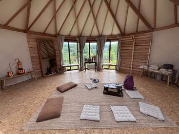 Una sala de meditación circular y tranquila, con grandes ventanales, madera clara y tonos neutros. Los cojines y las esterillas forman un círculo; los instrumentos y la decoración crean un ambiente apacible y acogedor.