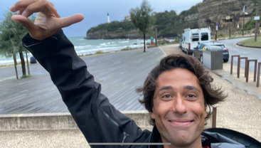 Ein Mann lächelt, während er draußen in Küstennähe ein Selfie macht. Er zeigt nach oben; hinter ihm sind das Meer und ein Leuchtturm in der Ferne zu sehen, der Himmel ist bewölkt.