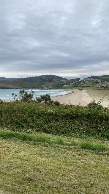 Ein bewölkter Himmel überzieht eine friedliche Küstenlandschaft mit sanften Hügeln, Sandstrand, sanften Wellen, grünem Gras, Sträuchern, vereinzelten Häusern und einer ruhigen, ländlichen Dorfatmosphäre.