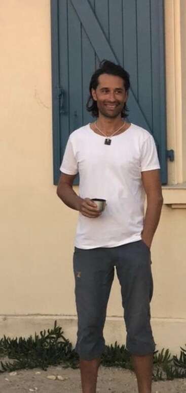 Ein lächelnder Mann steht draußen vor einer beigen Wand und blauen Fensterläden und hält eine Tasse in der Hand. Er trägt ein weißes T-Shirt, eine graue Hose und eine Halskette und wirkt ganz entspannt.