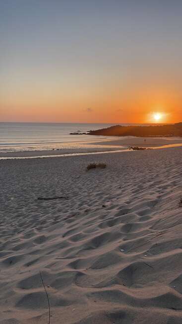 Sanftes, orangefarbenes Sonnenlicht taucht über einem ruhigen Strand unter und wirft goldene Reflexe auf die sanften Wellen. Sanfte Sanddünen und Silhouetten in der Ferne schaffen eine friedliche, beschauliche Atmosphäre.