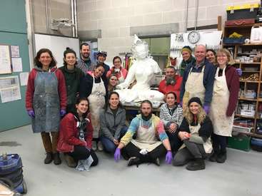 The image depicts a group of people posing for a photo in an art studio, with a large white sculpture of a woman in the background.
