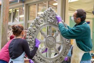 The image shows a group of people working on a large piece of artwork, possibly a sculpture, with a table and tools visible in the background. The scene appears to be a collaborative effort.
