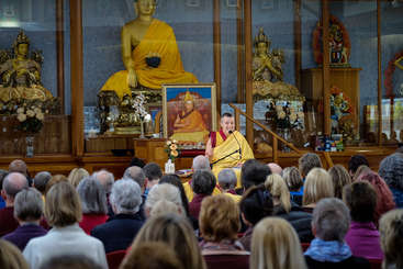 The image depicts a serene scene of a Buddhist monk speaking to a group of people, with a crowd of onlookers seated in front of him. The atmosphere is peaceful and contemplative.