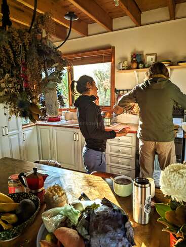 Dos personas preparan comida en una cocina acogedora e iluminada por el sol. La encimera y la mesa muestran frutas, verduras, una cafetera y un jarrón con flores.