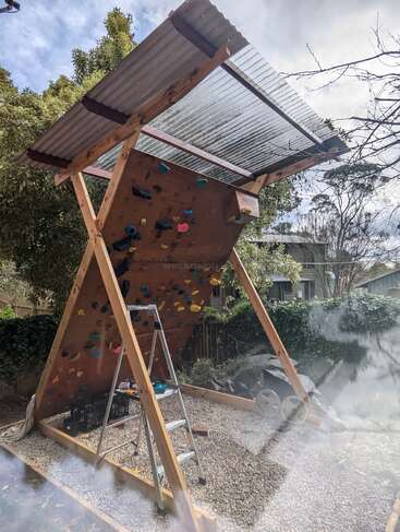 Esta imagen muestra un rocódromo casero al aire libre bajo un techo ondulado transparente. Hay una escalera metálica, una base de grava, cajas y árboles al fondo.