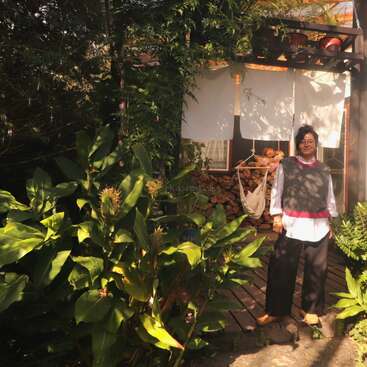 A woman stands in front of a cozy wooden house surrounded by lush green plants. Firewood is stacked beside her, sunlight and shadows fill the scene.