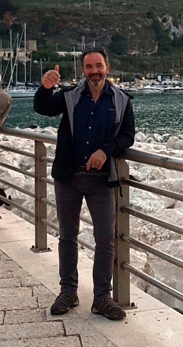 Um homem sorridente está em pé perto de uma grade à beira-mar, fazendo um sinal de positivo com o polegar. Ele está vestido com roupas casuais, com os óculos de sol na cabeça, tendo barcos e montanhas ao fundo.