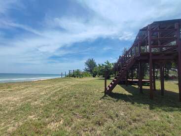 A coastal scene with grassy land, a wooden deck structure with stairs, some trees, and the beach meeting the blue ocean under a partly cloudy sky.