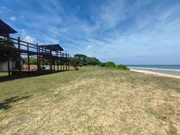 A wooden structure overlooks a sandy grassy area leading to a tranquil beach. Blue sky, scattered clouds, gentle waves, and distant greenery create a peaceful, scenic atmosphere.