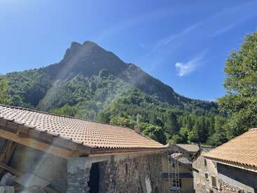 Dieses Bild zeigt im Vordergrund rustikale Steinhäuser mit Ziegeldächern, einen üppig grünen Wald und einen hoch aufragenden Berggipfel unter einem strahlend blauen Himmel.