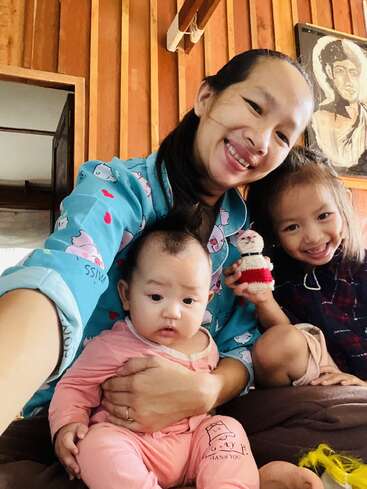 A smiling woman sits indoors with her two young children, one baby in pink pajamas and an older girl holding a toy, capturing a joyful family moment.