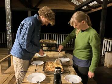 Two people prepare to share a meal outside at night, serving food from a large baking dish onto plates on a rustic wooden table under a shelter.