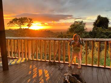 A child stands barefoot on a wooden deck at sunset, smiling and making peace signs. Vibrant orange sky and lush green trees create a beautiful, serene scene.
