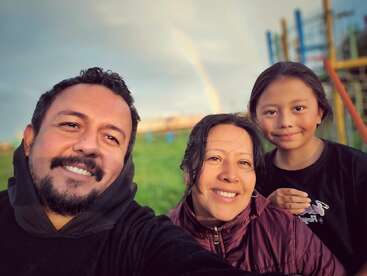 A cheerful family of three smiles outdoors. There's a playground and a vivid green field behind them, with a faint rainbow visible in the background sky.