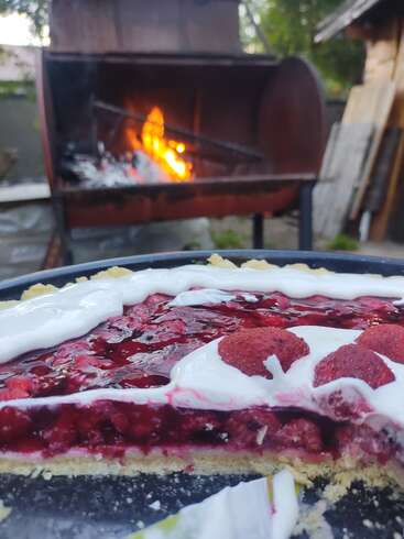 A delicious berry pie topped with cream sits in the foreground, while a barbecue with a glowing fire blazes in the background, outdoors in a rustic setting.