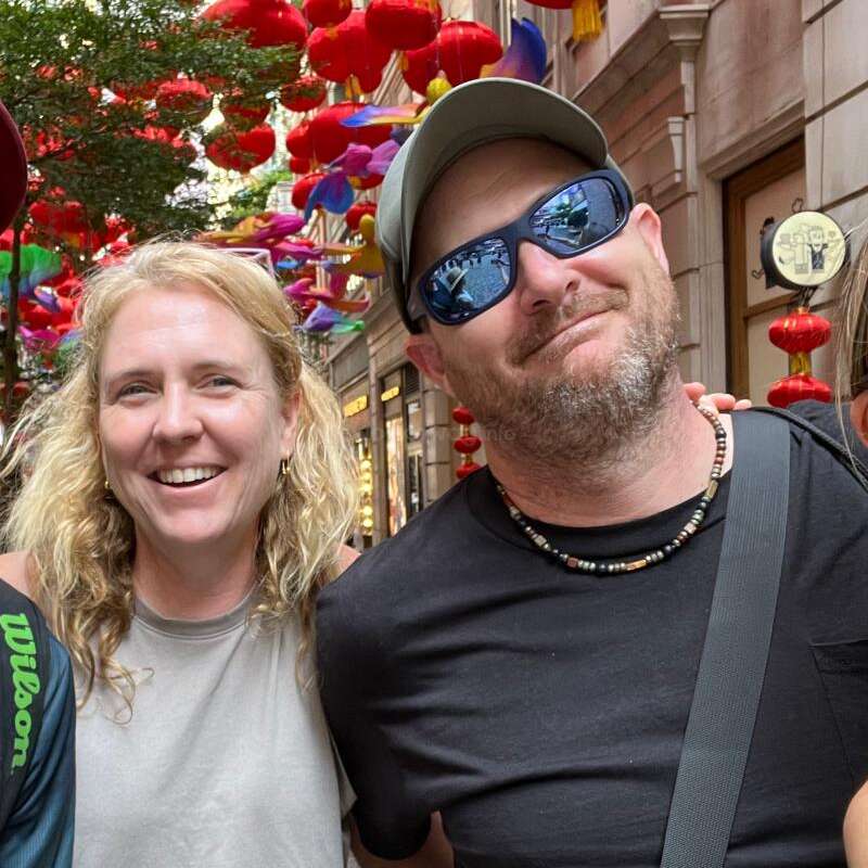 Un homme et une femme souriants posent ensemble en plein air, entourés de lanternes rouges vibrantes et de décorations colorées suspendues au-dessus d'une rue animée de la ville, dégageant une énergie joyeuse.
