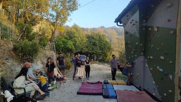 Un groupe de personnes se réunit en plein air près d'une zone boisée, certaines assises et discutant, d'autres regardant une femme escalader un mur d'escalade avec des crash pads en dessous.