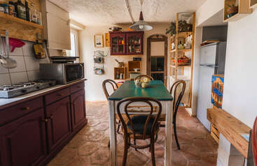 Cette cuisine confortable présente un décor rustique, des meubles en bois, une table à plateau vert avec des chaises, des étagères avec divers objets, une cuisinière, un réfrigérateur et un éclairage naturel chaleureux.
