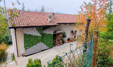 Une maison blanche au toit de tuiles est dotée d'un mur d'escalade extérieur sur un de ses côtés. Du bois est soigneusement empilé en dessous. Des arbres au feuillage d'automne entourent la maison.